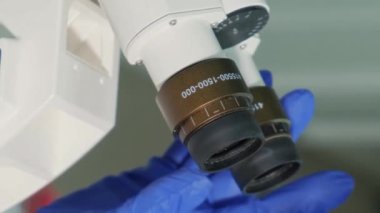 Kadın bir laboratuvar asistanı mikroskop yardımıyla maske, gözlük ve tıbbi kapak takarak virüsü inceliyor. Dikey video.