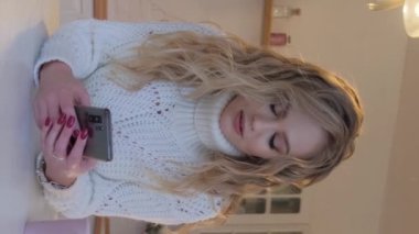 Beyaz kazaklı güzel sarışın bir kadın akıllı telefona bakıyor ve gülümsüyor. Dikey video.