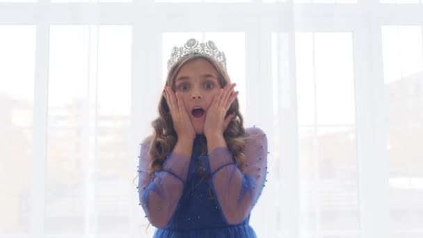 드레스와 왕관에있는 소녀는 놀라게된다 창문과 소파의 배경에 아이의 클로즈업 초상화 — 비디오