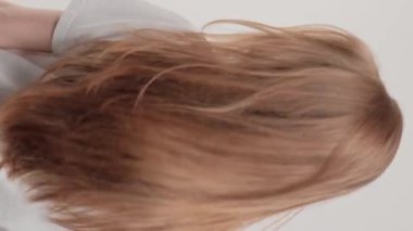 Dikey video. Kızıl saçlı bir kız sırtı gri bir arka planda stüdyoda duruyor ve saçını sallıyor. Güzel ve sağlıklı saçlarla ilgilenme konsepti.