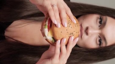 Dikey video. Güzel esmer, sulu bir burger yiyor, yakın plan portre..