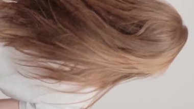 Dikey video. Kızıl saçlı bir kız sırtı gri bir arka planda stüdyoda duruyor ve saçını sallıyor. Güzel ve sağlıklı saçlarla ilgilenme konsepti.