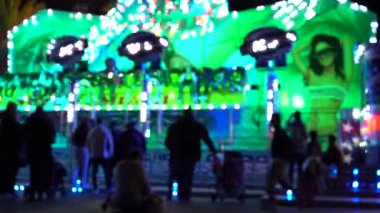 Eğlence parkındaki atlıkarıncaların renkli ışık hareketleriyle ve dönme dolabın arka planıyla gece manzarası. Bulanık video.