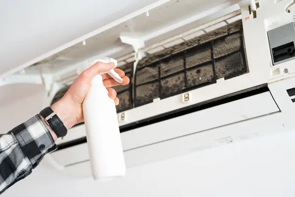 A mans hand sprays a cleaning liquid on the moldy filters of an air conditioner.