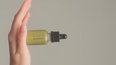 Güzel kadınların elleri şeffaf beyaz bir şişede kozmetik yağı tutar, gri arka planda. Dikey video.