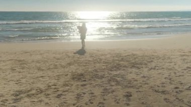 Sahilde küçük bir kız denizin arka planında beyaz küçük bir köpekle koşuyor..