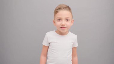 Beyaz tişörtlü küçük bir çocuk düşünceli bir şekilde kameraya bakar ve sonra mutlu bir şekilde gülümser. Stop motion gri arka planda stüdyoda çekilmiştir.