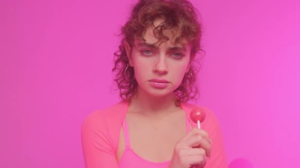 迷人美丽的年轻女子模特儿在粉红的背景上 吃着软糖棒棒糖 女孩舔糖果 — 图库视频影像