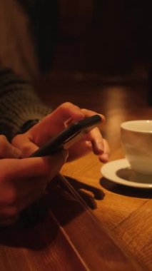 Barda elinde bir fincan kahveyle oturan bir adam akıllı telefonuna SMS yazıyor. Dikey video.