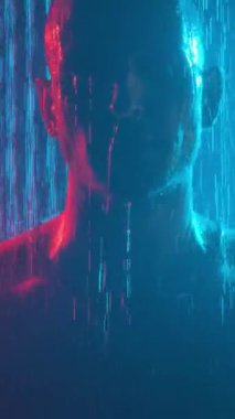 Portrait of a young pumped-up guy in the shower with fog in neon light on a black background. Vertical video.