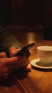 Barda elinde bir fincan kahveyle oturan bir adam akıllı telefonuna SMS yazıyor. Dikey video.