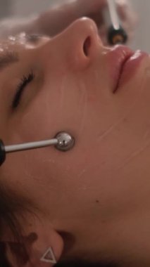 Bir güzellik uzmanının cilt bakımı prosedürleriyle yüzleşmek. Genç ve güzel bir kızın yüzünün mikro-akım terapisi. Dikey video.