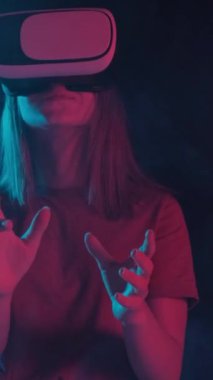 Gözlüklü ve tişörtlü bir kız neon ışıkta sanal gerçeklik oynuyor. Dikey video.