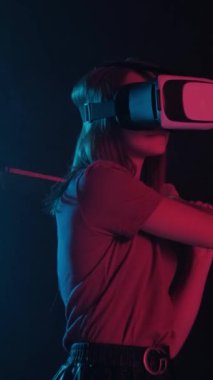 Gözlüklü ve tişörtlü bir kız neon ışıkta sanal gerçeklik oynuyor. Dikey video.