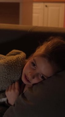 Şirin bir kız kanepenin üzerinde battaniyeyle televizyon karşısında uyuyakalıyor. Dikey video.