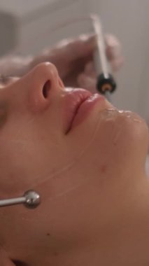 Bir güzellik uzmanının cilt bakımı prosedürleriyle yüzleşmek. Genç ve güzel bir kızın yüzünün mikro-akım terapisi. Dikey video.