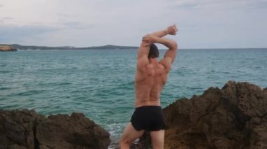 Şortlu bir adam, bir atlet, kayalıklarda ısınıyor denizin arka planında.