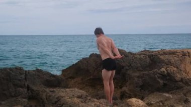 Şortlu bir adam, bir atlet, kayalıklarda ısınıyor denizin arka planında.