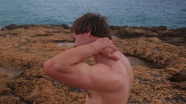 Şortlu bir adam, bir atlet, kayalıklarda ısınıyor denizin arka planında.