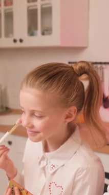 Evdeki mutfakta küçük bir kızın portresi. Çocuk dudaklarını pembe ruj ve göz kırparak boyuyor. Dikey video.