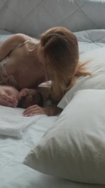 Genç anne, beyaz bir odada bebeğini yatakta emziriyor. Dikey video.