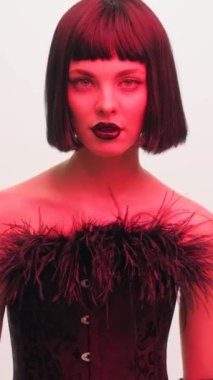 Fashion portrait of a girl in a black bob wig and black lipstick in red light on a white background. Black leather gloves. The girl shows the fuck. Vertical video.