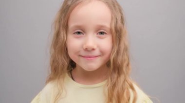 Küçük kız kameraya bakıp gülüyor. Stüdyodaki bir çocuğun portresi. Gri arka planda, yakın planda. Süper yavaş çekim.