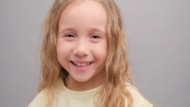Küçük kız kameraya bakıp gülüyor. Stüdyodaki bir çocuğun portresi. Gri arka planda, yakın planda. Süper yavaş çekim.