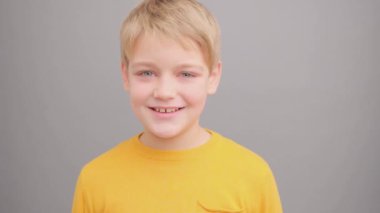 Sarı tişörtlü bir çocuk gri arka planda gülümsüyor ve kameraya bakarken sinirlenmeye başlıyor..