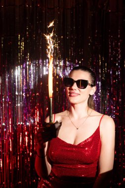 Çarpıcı kırmızı bir elbise giyen şık bir kadın havai fişekle kutlama yaparken canlı bir şenlik atmosferinin tadını çıkarıyor.