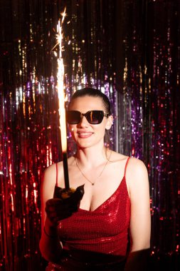 Çarpıcı kırmızı bir elbise giyen şık bir kadın havai fişekle kutlama yaparken canlı bir şenlik atmosferinin tadını çıkarıyor.