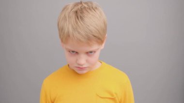 Sarı tişörtlü, gri arka planlı çocuk sinirlenir ve gülümser.