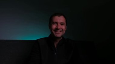Bir adam kameraya bakar ve güler, karanlık bir arka planı olan bir odada..