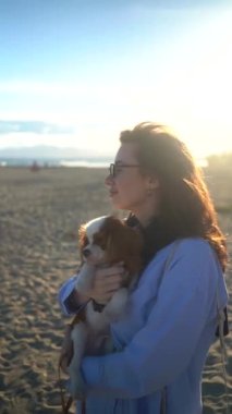 Genç bir kadın gün batımında kumsalda şövalyesi Kral Charles Spaniel 'e sarılıyor. Sıcak ışık, dalgalar ve gülümsemeler huzurlu, neşeli bir bağ ve rahatlama anını yakalar..