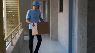 Açık mavi üniformalı ve şapkalı bir erkek kurye apartman kapısının önünde duran bir paket teslim ediyor..