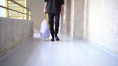 Bir kişi koridorda yürürken görülüyor, bir elinde büyük bir çöp torbası, siyah pantolon ve terlik giymiş..