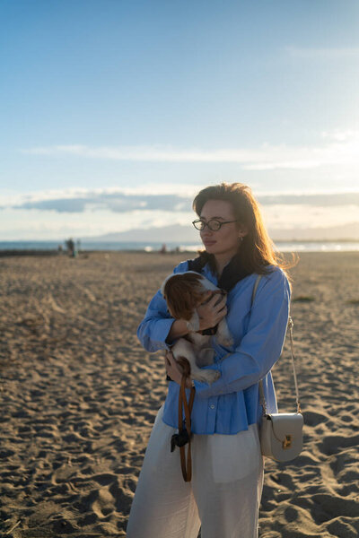 Молодая женщина обнимает своего короля Кавалера Чарльза Спаниеля на пляже на закате. Теплый свет, волны и улыбки захватывают мирный, радостный момент общения и релаксации.