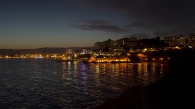 Sakin deniz suyuna yansıyan aydınlık binalarla dolu bir sahil şehrinin gece görüntüsü günbatımından hemen sonra sakin ve renkli bir atmosfer yaratıyor..