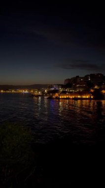 Sakin deniz suyuna yansıyan aydınlık binalarla dolu bir sahil şehrinin gece görüntüsü günbatımından hemen sonra sakin ve renkli bir atmosfer yaratıyor..