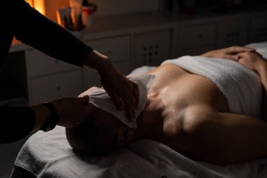 Bir adam masaj masasında havluyla kaplıyken yüz spa tedavisi görüyor. Bir profesyonel nazikçe yüzüne havlu yapıştırır ya da sıkıştırır. Atmosfer sakin ve rahatlatıcı, sıcak, yumuşak ışıklandırmayla.