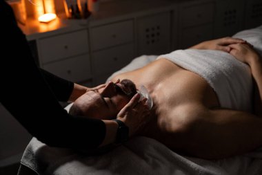 Bir adam masaj masasında havluyla kaplıyken yüz spa tedavisi görüyor. Bir profesyonel nazikçe yüzüne havlu yapıştırır ya da sıkıştırır. Atmosfer sakin ve rahatlatıcı, sıcak, yumuşak ışıklandırmayla.