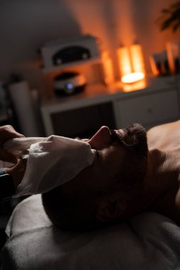 Bir adam masaj masasında havluyla kaplıyken yüz spa tedavisi görüyor. Bir profesyonel nazikçe yüzüne havlu yapıştırır ya da sıkıştırır. Atmosfer sakin ve rahatlatıcı, sıcak, yumuşak ışıklandırmayla.