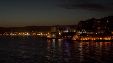 Sakin deniz suyuna yansıyan aydınlık binalarla dolu bir sahil şehrinin gece görüntüsü günbatımından hemen sonra sakin ve renkli bir atmosfer yaratıyor..