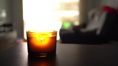 Karanlık bir yüzeyde, cam bir kavanozda yanan bir mum, arka plandaki sıcak, bulanık bir iç mekanı hafifçe aydınlatıyor..
