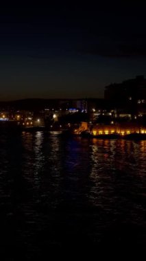 Sakin deniz suyuna yansıyan aydınlık binalarla dolu bir sahil şehrinin gece görüntüsü günbatımından hemen sonra sakin ve renkli bir atmosfer yaratıyor..