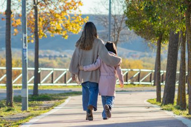 Bir anne ve kızının, sonbahar ağaçlarıyla çevrili huzurlu bir yolda birlikte yürüdükleri yürek ısıtan bir sahne. Her ikisi de rahat bir sonbahar paltosu giymiş, manzarayla dolu, huzurlu bir yürüyüşün tadını çıkarıyorlar..