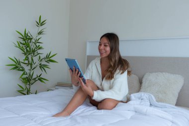 pretty latina woman on a white bed with soft cushions in a quiet room using a tablet relaxing and enjoying a relaxing moment with a green plant in the background, and a beige headboard in a quiet and clipart