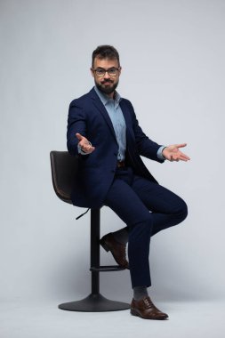 Tabureye yan oturmuş, avuçları yukarı bakan ve sakin bir ifadeyle kolları açık İspanyol bir adamın zarif tam vücut fotoğrafı. Güçlü bir poziyon, açıklık, duygusal açıklık ve güç..