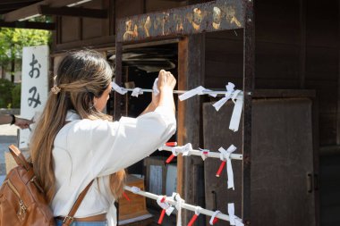 Genç bir yolcunun dileğini Japon tapınağındaki ahşap bir yapıya bağlaması, beyaz gömlek ve kahverengi sırt çantası giymesi gelenek ve kültürel deneyimi sembolize ediyor.