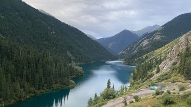 Kolsay Gölü yüksek yeşil dağların eteklerinde mavi suyla. Su sakin, çam ağaçları uzun ve gökyüzü gri..